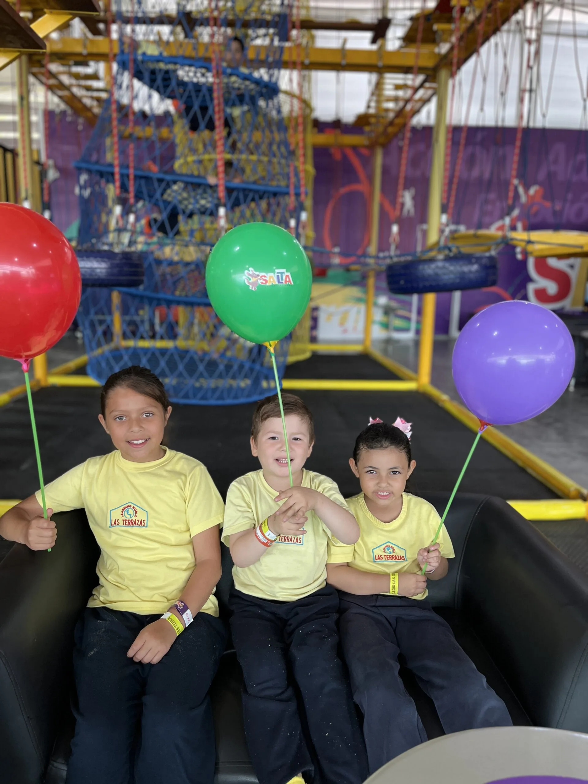Tres niños del Preescolar Las Terrazas sonriendo con globos de colores en un área de juegos recreativa.